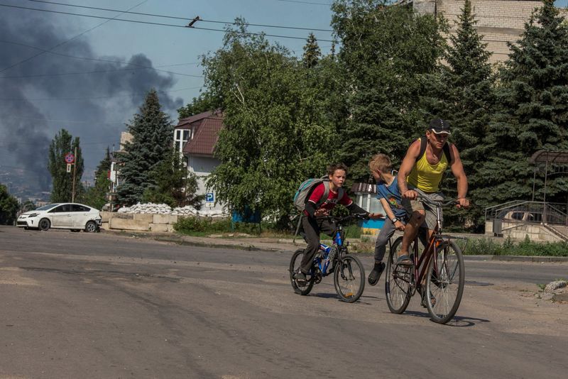 © Reuters. A local man with kids rides a bicycle along an empty street, as smoke rises after shelling in the background, amid Russia's attack on Ukraine, in the town of Lysychansk, Luhansk region, Ukraine June 10, 2022. REUTERS/Oleksandr Ratushniak