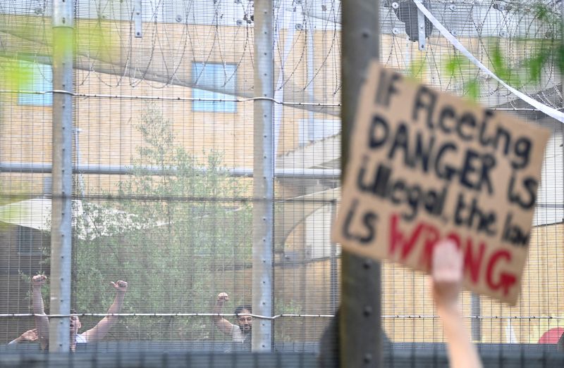 &copy; Reuters. Varias personas detenidas en un centro de inmigrantes hacen gestos hacia un grupo que protesta en contra de la deportación de solicitantes de asilo a Ruanda, uno de cuyos participantes porta un cartel que dice en inglés "Si huir del peligro es ilegal, l