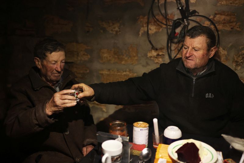 © Reuters. Stepan Kovalyov, 80, and his brother Volodymyr Kovalyov, 77, toast with horilka, a Ukrainian spirit, inside of a cellar where Stepan and his wife live after their house was destroyed during the Russian occupation, amid Russia's invasion of Ukraine, in the village of Posad-Pokrovske, northwest of the city of Kherson, Ukraine January 29, 2023. There is no central power or heating and the surrounding fields are heavily mined, making them unworkable. Yet the brothers have decided to stay in the isolated farming village to live out their days in the place they know best.      REUTERS/Nacho Doce  