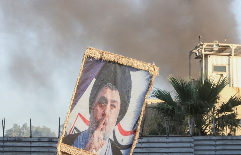 © Reuters. A protester holds up a portrait of Iraqi Shi'ite cleric Moqtada al-Sadr as smoke rises from the Swedish embassy building in Baghdad during a protest near the embassy hours after it was stormed and set on fire ahead of an expected Koran burning in Stockholm, in Baghdad, Iraq, July 20, 2023. REUTERS/Ahmed Saad
