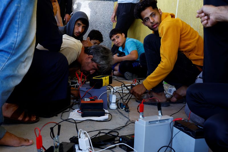© Reuters. FILE PHOTO: Palestinians charge their mobile phones from a point powered by solar panels provided by Adel Shaheen, an owner of an electric appliances shop, as electricity remains cut during the ongoing Israeli-Palestinian conflict, in Khan Younis in the southern Gaza Strip October 19, 2023. REUTERS/Mohammed Salem