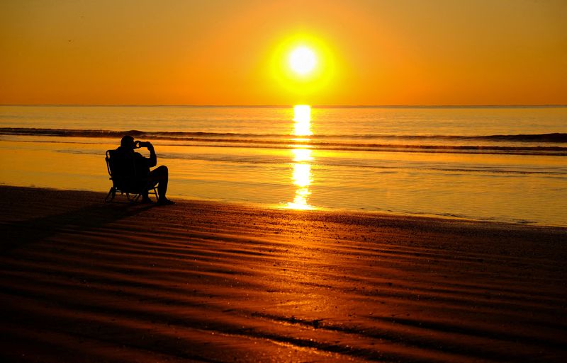 &copy; Reuters. FILE PHOTO: Sitting on a beach at water?s edge, a man takes a photo as the sun rises over Hilton Head Island, South Carolina, U.S., March 16, 2023. REUTERS/Kevin Lamarque/File Photo