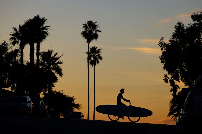 © Reuters. FILE PHOTO: A surfer carries his surfboard on his bicycle as his pedals his way from the beach after surfing in Encinitas, California, U.S., January 9, 2024.  REUTERS/Mike Blake/File Photo