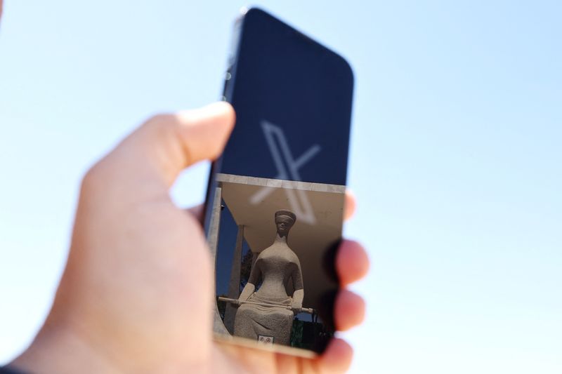 © Reuters. The logo of social media platform X is seen on a mobile phone next to a reflection of Alfredo Ceschiatti's sculpture The Justice, outside Brazil's Supreme Court in this illustration taken August 30, 2024, in Brasilia, Brazil.   REUTERS/Ueslei Marcelino