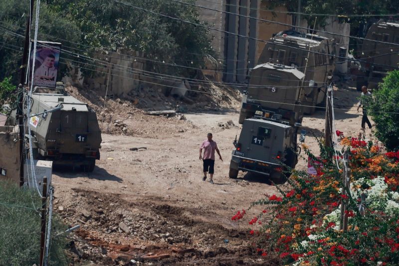 © Reuters. A Palestinian man walks near Israeli military vehicles, during an Israeli raid in Jenin, in the Israeli-occupied West Bank, August 31, 2024. REUTERS/Ammar Awad