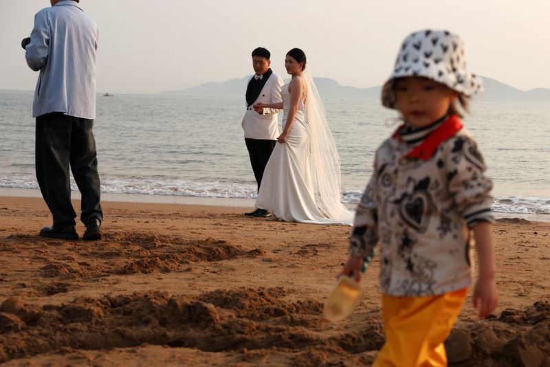 © Reuters. FILE PHOTO: A child plays with sand near a couple taking part in a pre-wedding photoshoot on a beach in Qingdao, Shandong province, China April 21, 2024. REUTERS/Florence Lo/File Photo