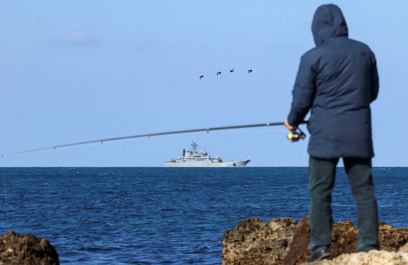 © Reuters. FILE PHOTO: A man fishes on the shore as an amphibious assault ship of the Russian Navy sails near the Black Sea port of Sevastopol, Crimea February 16, 2022. REUTERS/Alexey Pavlishak/File Photo