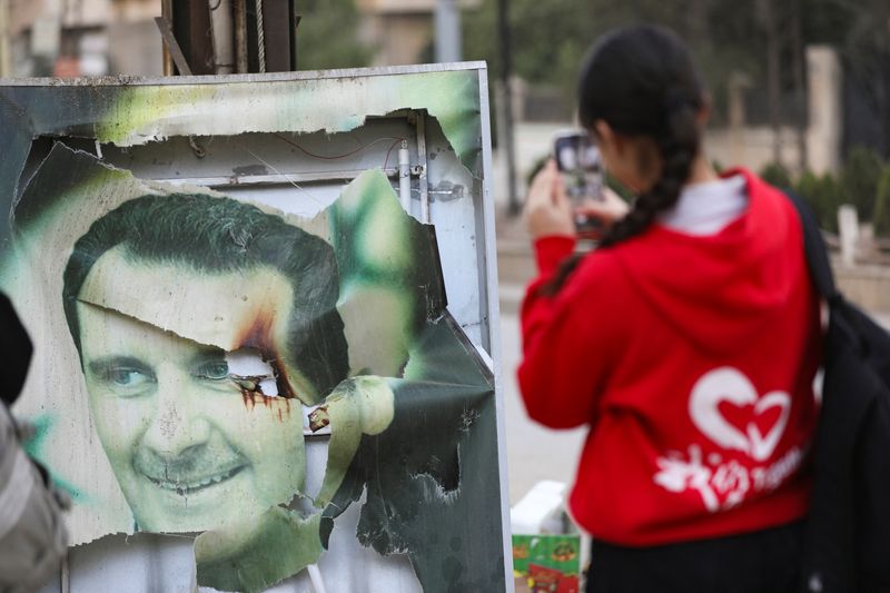 © Reuters. A woman uses her cellphone near a damaged photo of Syrian President Bashar al-Assad as people celebrate, after Syrian rebels announced they had ousted President Bashar al-Assad, in Qamishli, Syria, December 8, 2024. REUTERS/Orhan Qereman