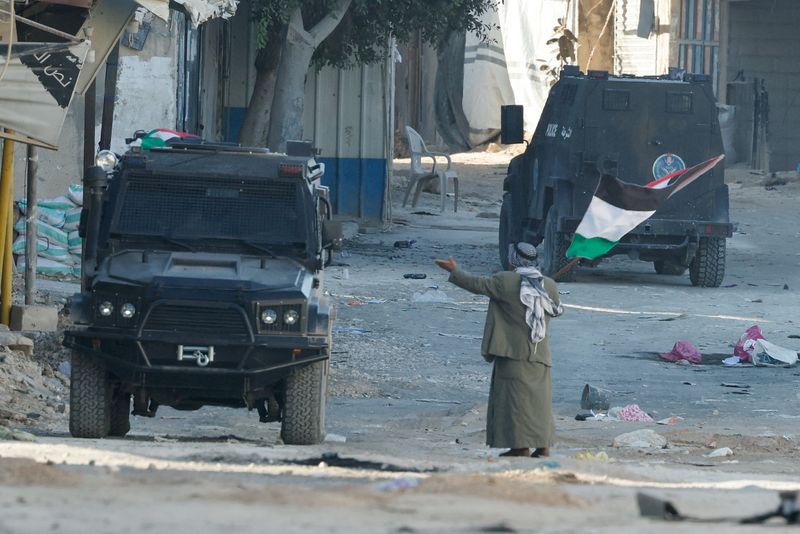 © Reuters. A man holding a Palestinian flag gestures next to a vehicle of Palestinian security forces amid clashes with militants at the camp, in Jenin in the Israeli-occupied West Bank, December 14, 2024. REUTERS/Raneen Sawafta