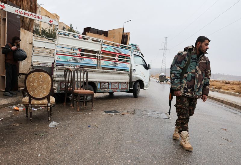 © Reuters. A (HTS) fighter and the Syrian ruling body walks alongside workers putting the belongings of the families of former soldiers and officers of Assad's regime army out of the apartment on a truck as they leave their homes in the officer housing on the outskirts of Damascus following evacuation orders from factions of Hay'at Tahrir al-Sham (HTS), after Syria's Bashar al-Assad was ousted, in Syria, December 29, 2024. REUTERS/Amr Abdallah Dalsh