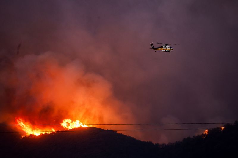 © Reuters. A helicopter flies as smoke and fire rise from the Palisades Fire threatening homes in the Brentwood area of ​​Los Angeles, California, US, January 11, 2025. REUTERS/Shannon Stapleton