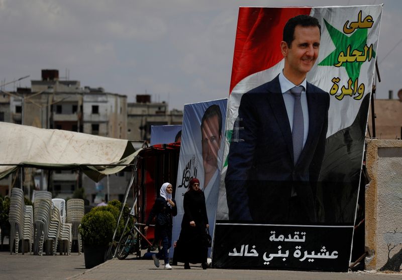 &copy; Reuters. امرأتان تمران عبر لافتة انتخابية للرئيس السوري بشار الأسد في حمص يوم 23 مايو آيار 2021. تصوير: عمر صناديقي - رويترز.