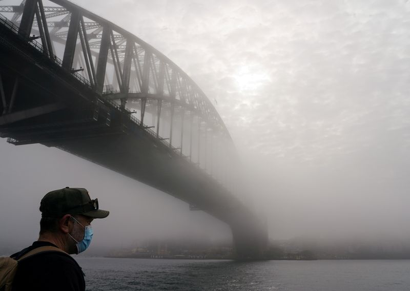 © Reuters. FILE PHOTO: A man wearing a protective face mask takes in the waterfront view under the Sydney Harbour Bridge, seen shrouded in morning fog, during a lockdown to curb the spread of a coronavirus disease (COVID-19) outbreak in Sydney, Australia, July 1, 2021. REUTERS/Loren Elliott/File Photo