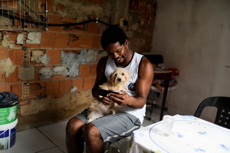 &copy; Reuters. Felipe Luther, 37, a scholarship student at Pontifical Catholic University of Rio (PUC-Rio) who also works as a street-sweeper, uses his cellphone to take part in an online class for his university, as he holds his dog Mel at his home in Belford Roxo, nea