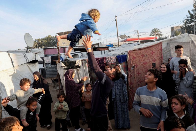 © Reuters. A man throws a child into the air as displaced Palestinians celebrate at a tent camp following a ceasefire between Israel and Hamas, in Deir Al-Balah in the central Gaza Strip, January 19, 2025. REUTERS/Ramadan Abed     